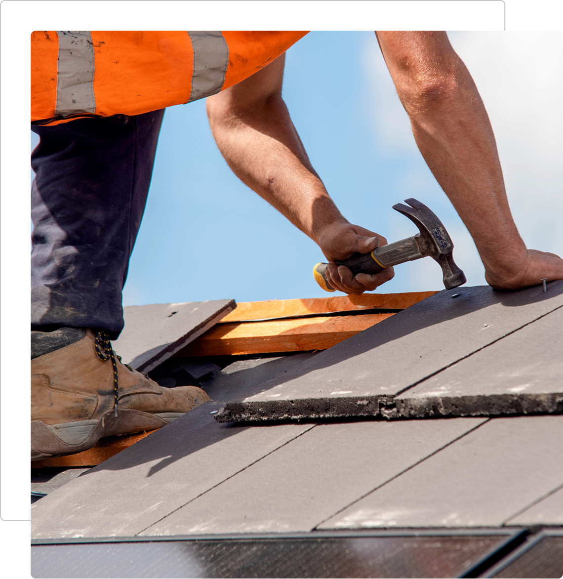 A worker hammering nails on a roof under clear blue sky.
