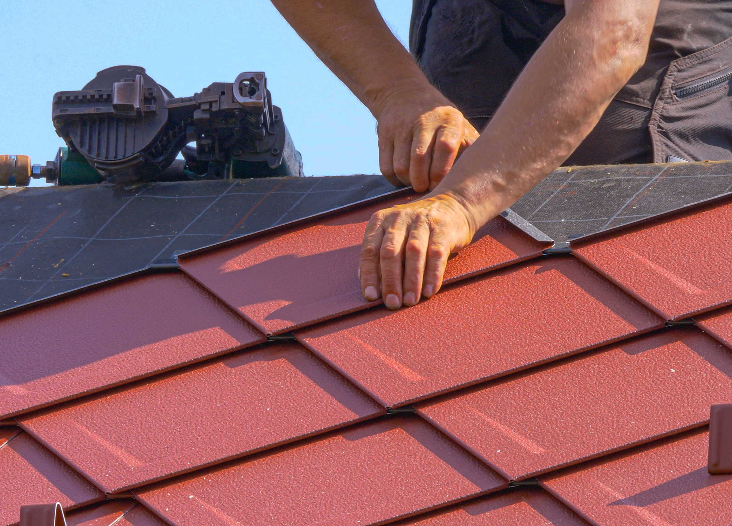 Hands installing red roof shingles on a sunny day.