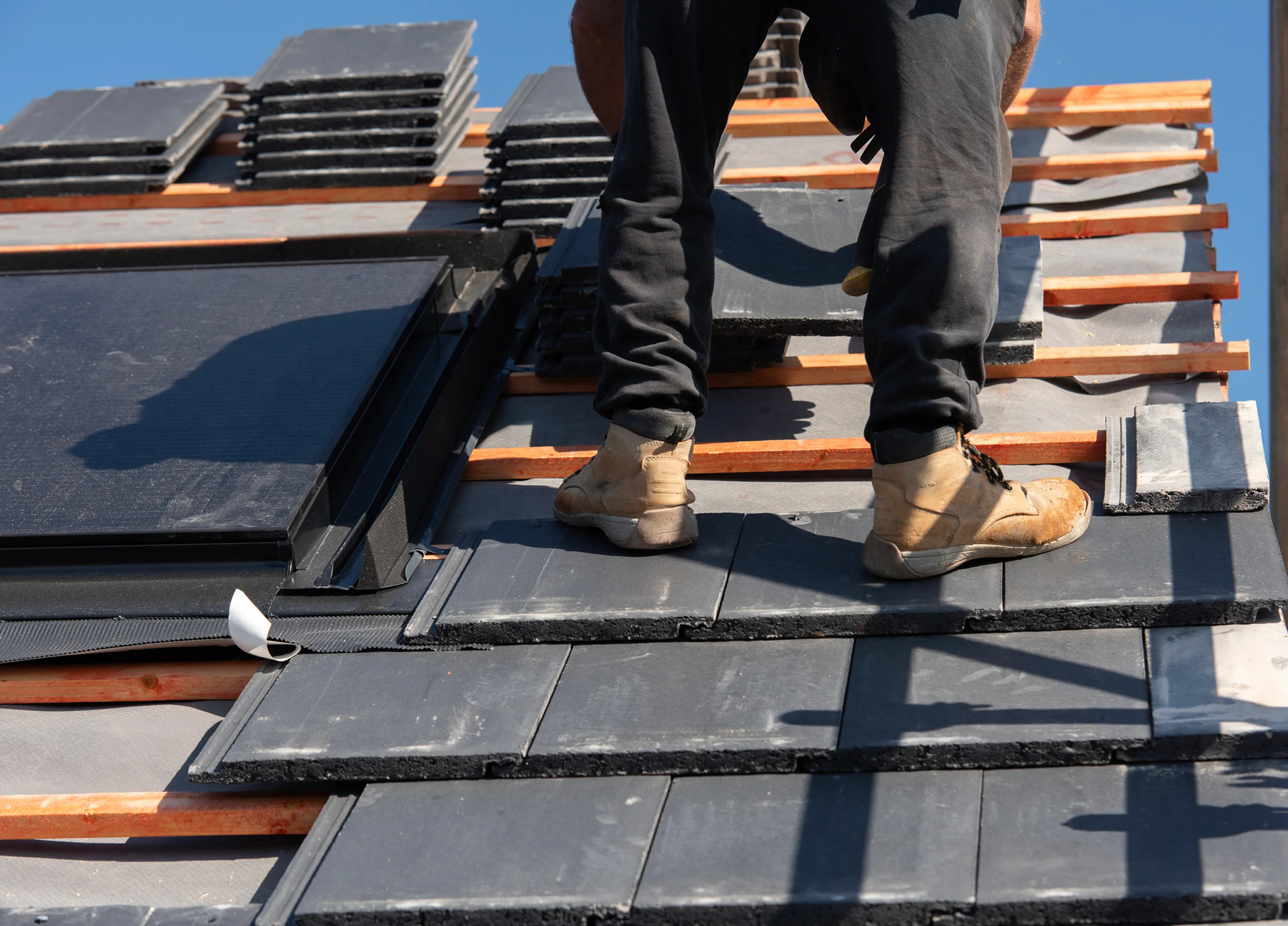 Person installing slate roof tiles with safety harness on a sunny day.